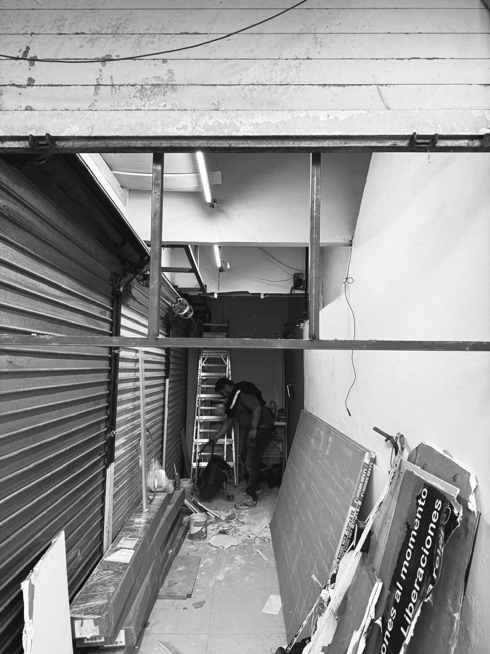Hombre trabajando en un local en remodelación rodeado de materiales de construcción y persianas metálicas.